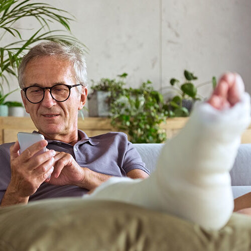 Cheerful man with broken leg in plaster cast sitting on sofa at home, life insurance